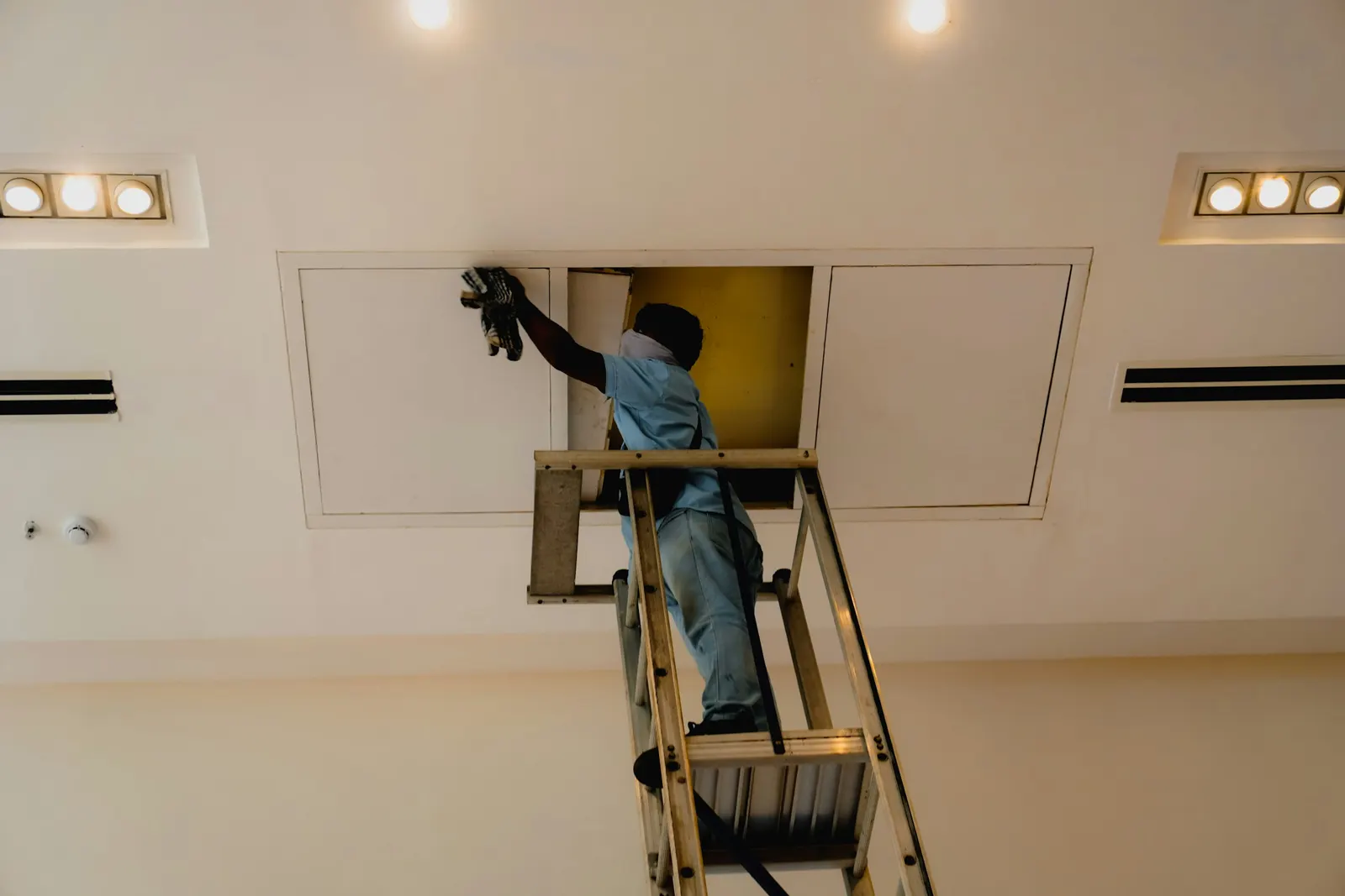 Worker on a ladder reaching into a ceiling access panel between air diffusers