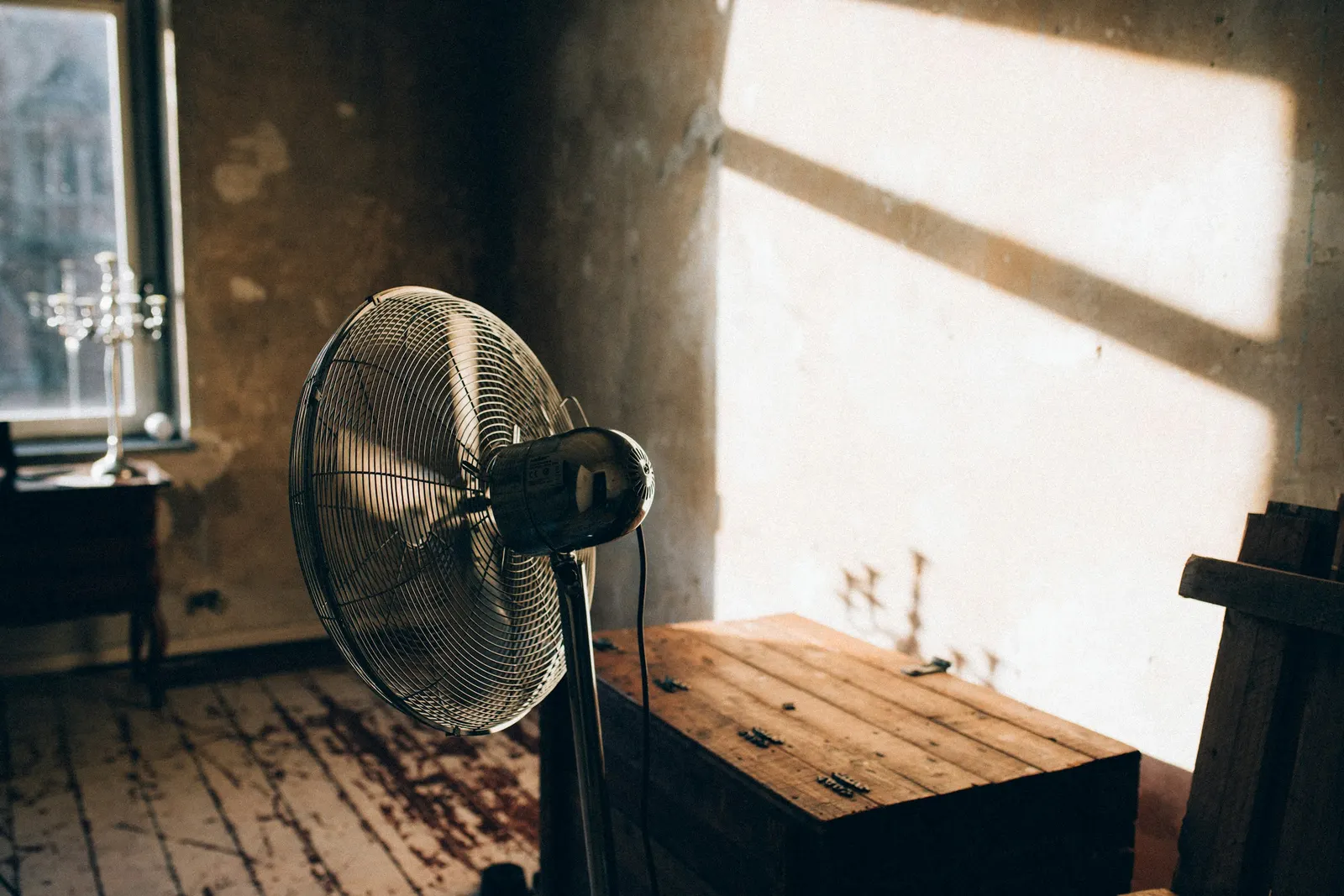 A pedestal fan in a warm, sunlit room with bare plaster walls.