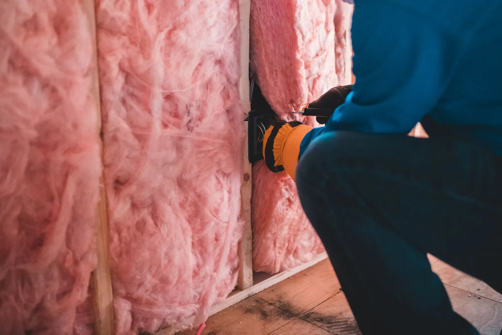 Worker in gloves fitting pink fiberglass insulation between wall studs