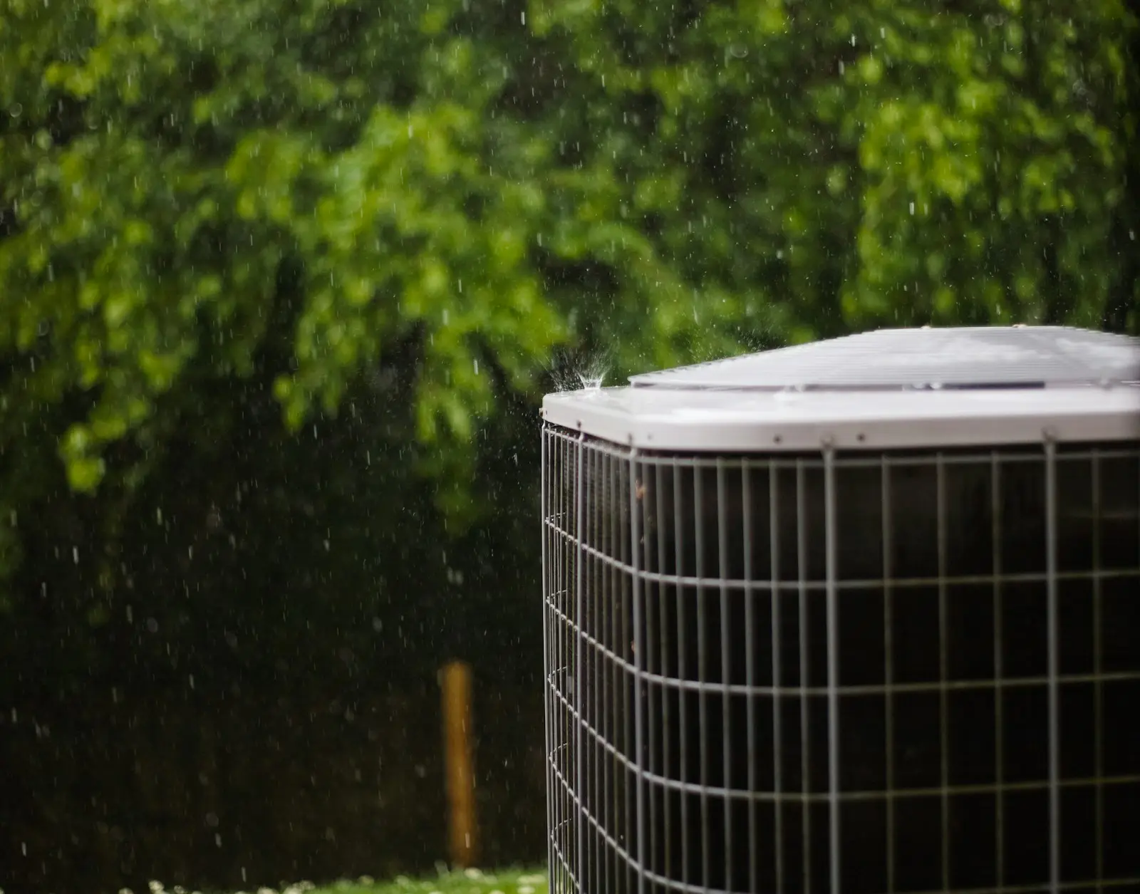 An outdoor central air conditioner unit in the rain, in front of green shrubs